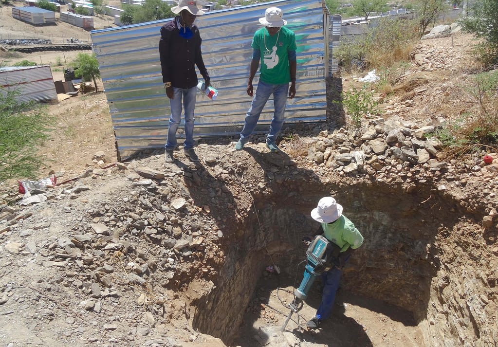 Two men watch as a third operates a drill in an earthen pit.