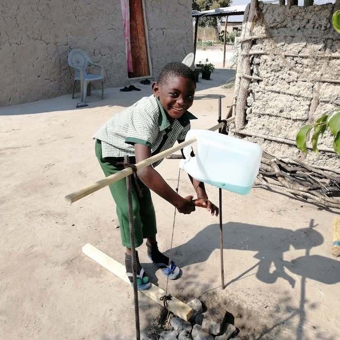A young boy operates a foot pedal controlled water source, known as a tippy-tap.