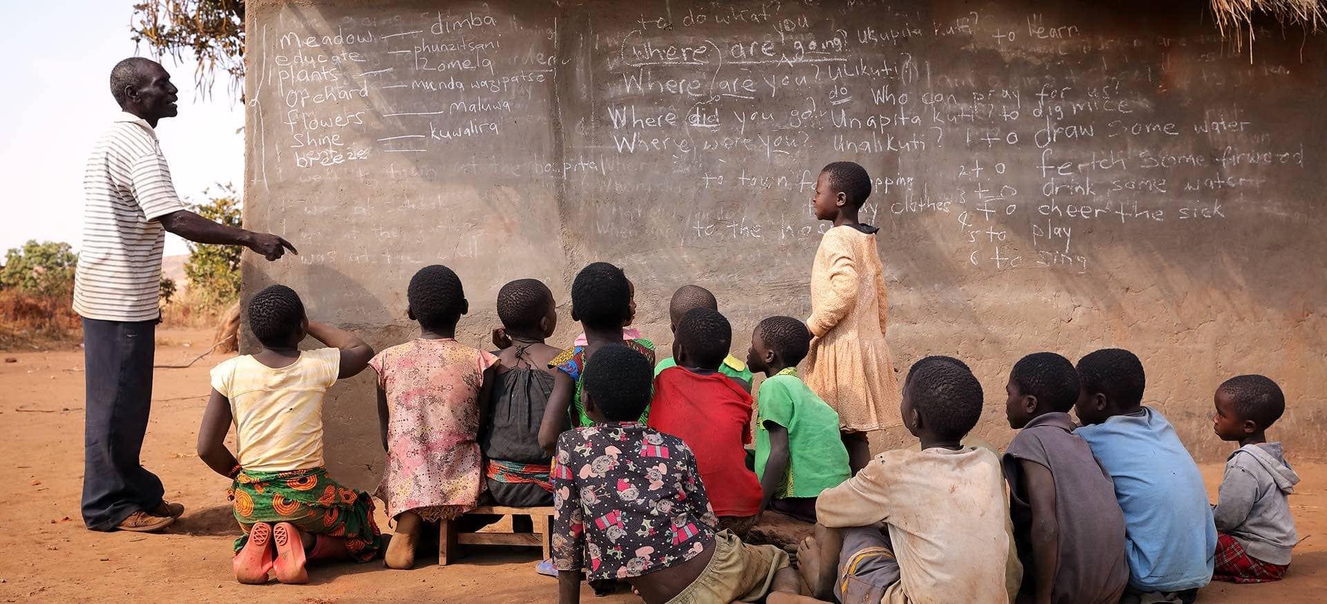 A teacher points towards a blackboard on the side of a hut while a group of children watch attentively.